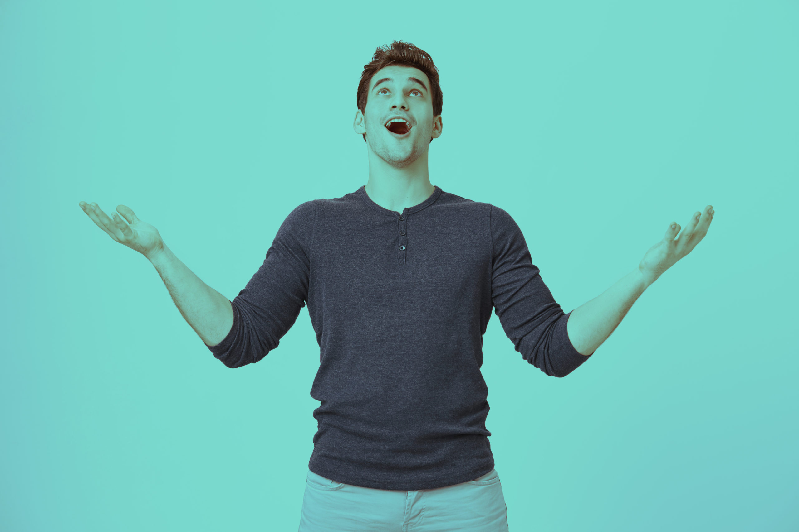 Portrait of fascinated excited young guy looking up with happy expression and raising arms, standing over gray background. Boyfriend sings to gilfriend while standing near her balcony.